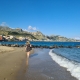 Frau mit Sonnenhut spaziert am Sandstrand von Giardini-Naxos entlang, im Hintergrund ein Küstenort und Berge auf Sizilien